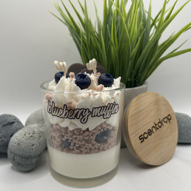 Glass container with layered dessert labeled 'blueberry muffin' next to a wooden lid with 'scented' branding, against a neutral background with plants and stones.