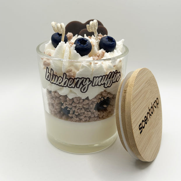 Scented candle in a glass jar with wooden lid labeled 'Blueberry Muffin' on a white background