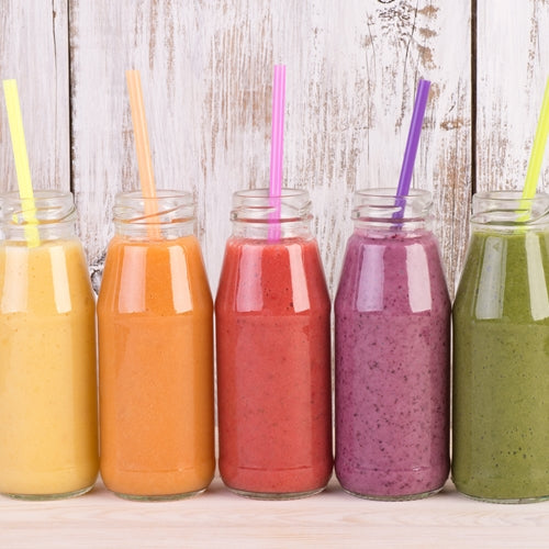 Five bottles of colorful smoothies with straws on a wooden surface.