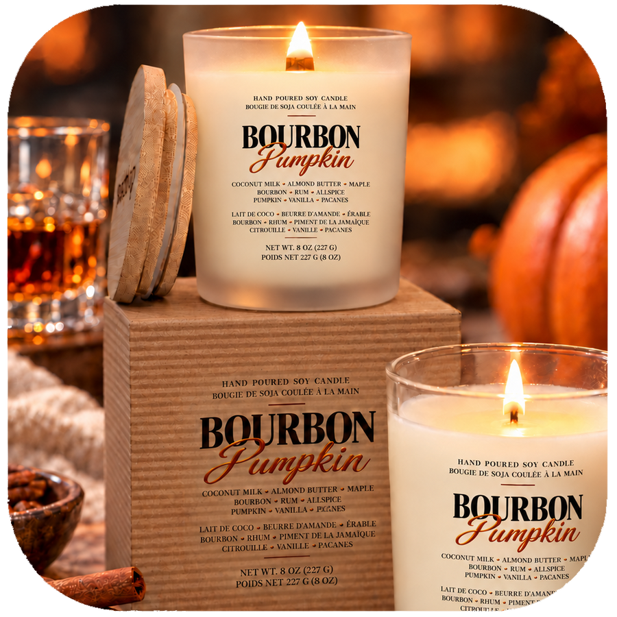 Two lit hand-poured soy candles labeled Bourbon Pumpkin are shown in a warm, cozy indoor setting with wooden coasters and a wooden box. The candles are white with black and orange text, and the background features warm, blurred amber tones.