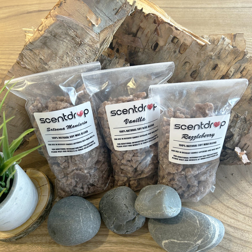 Three packages of Scentdrop wax melts on a wooden surface with rocks and a plant.