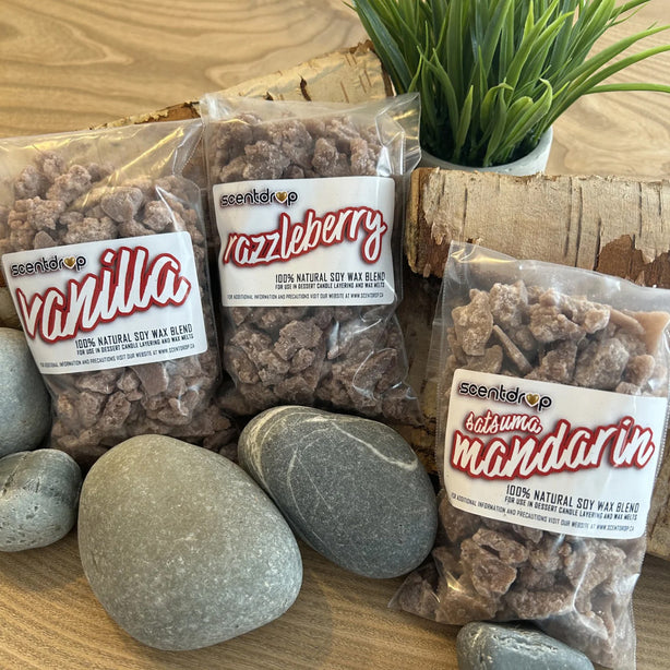 Three clear plastic bags of scentup natural soy wax blend pet chews arranged on a wooden surface with decorative stones and a small potted plant. The bags are labeled Vanilla, Nazzleberry, and Kalusha Mandarin.