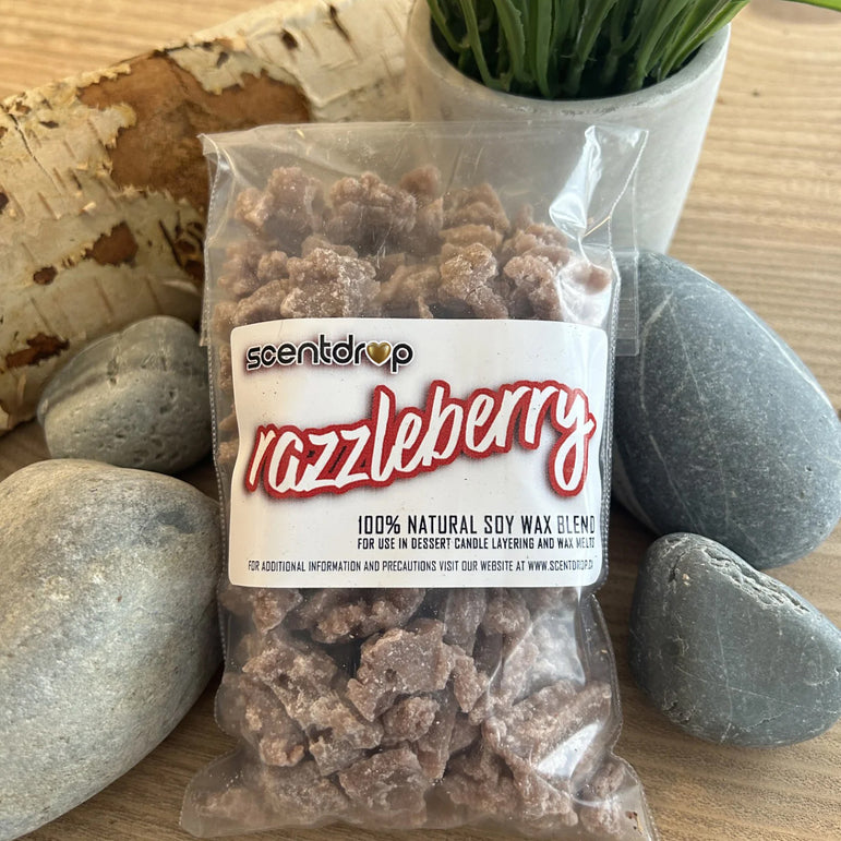 A clear plastic bag of scentdrop 'razzleberry' soy wax blend candles, filled with chunky brown wax pieces, displayed on a wooden surface surrounded by decorative stones and a small potted plant.