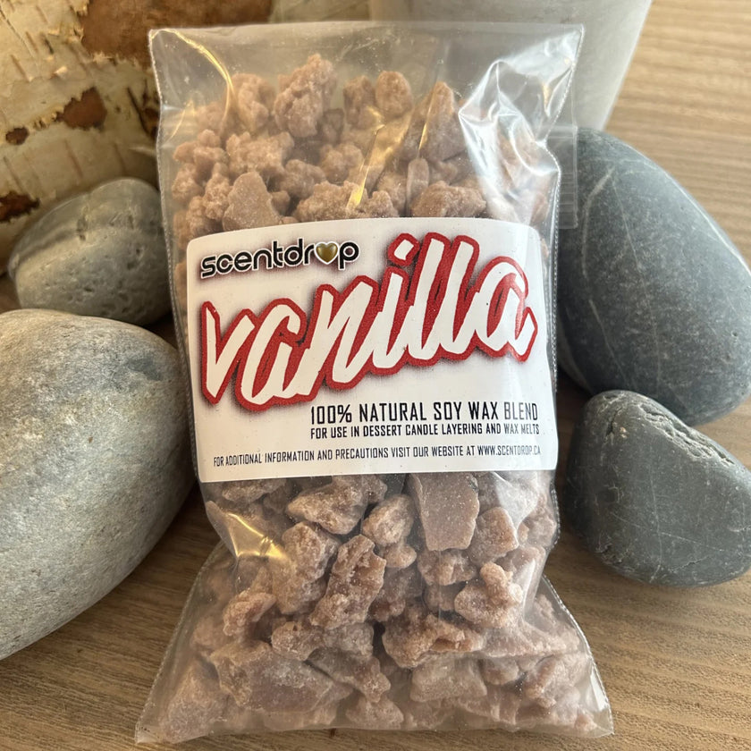 A clear plastic bag of Scentdrop Vanilla 100% natural soy wax blend, filled with chunky light brown wax pieces, displayed on a wooden surface surrounded by smooth gray stones.
