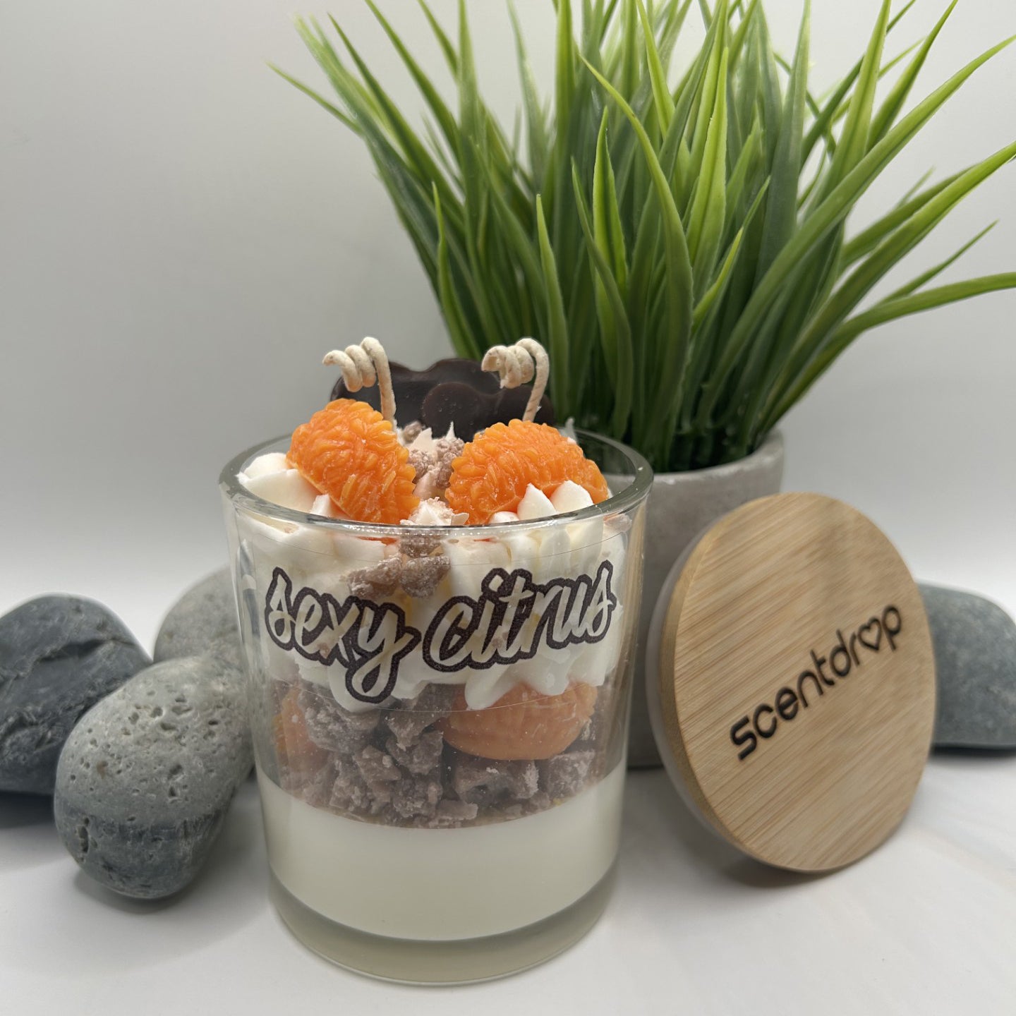 Scented candle in a glass jar with decorative elements and a wooden lid, set against a neutral background with plants and stones.